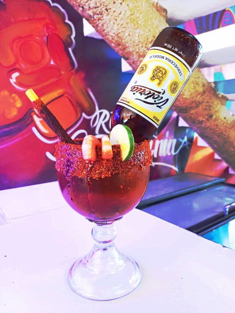 Una michelada o cerveza preparada con una botella de cerveza Victoria insertada, mariscos como camarones cocidos, limón y un popote con caramelo enchilado, servida en una copa escarchada con chile en polvo, sobre un fondo colorido y vibrante, ideal para disfrutar en un restaurante de marisco.